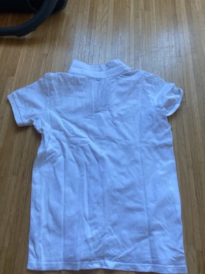 Vit pikétröja med krage - Klassisk vit pikétröja med korta ärmar och krage. Tröjan har knappslå framtill och en liten broderad logga på bröstet. Perfekt för en clean och sportig look.