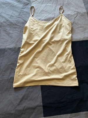 Beige linnetopp med smala axelband - Snygg beige linnetopp med smala, justerbara axelband och enkel design. Toppen har en liten broderad logga framtill och är tillverkad i ett mjukt, stretchigt material som känns skönt mot huden. Perfekt att styla med jeans eller kjol.