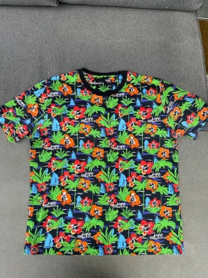 Färgglad blommig t-shirt Love Moschino - Säljer en färgglad t-shirt från Love Moschino med blommigt mönster i rött, orange, grönt och blått. T-shirten har svart rund halsringning och korta ärmar. Perfekt för dig som vill sticka ut med en unik och lekfull stil.
