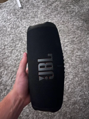 JBL svart bärbar högtalare - Snygg svart bärbar högtalare från JBL med robust design och tydlig logga på framsidan. Högtalaren har rundad form och är klädd i slitstarkt tygmaterial, perfekt för att ta med överallt och spela musik trådlöst.