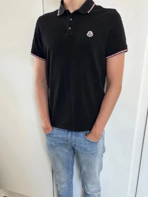 Moncler Svart pikétröja med rödvit kant - Moncler. Snygg svart pikétröja med krage och knappar framtill. Tröjan har röda och vita detaljer på krage och ärmslut samt en broderad logga på bröstet. Perfekt till jeans eller shorts för en clean look.