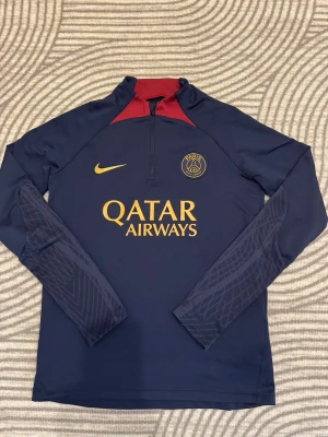 Mörkblå PSG träningströja Nike BARN 147-158cm - Snygg mörkblå långärmad träningströja från PSG med guld Nike-logga och Qatar Airways-tryck på bröstet. Tröjan har halv dragkedja, röda detaljer vid kragen och diskret mönster på ärmarna. Perfekt för fotbollsträning eller chill.