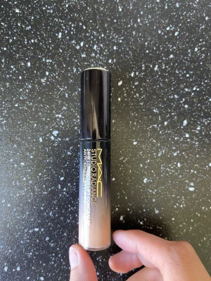 MAC Studio Radiance Concealer - MAC Studio Radiance Concealer med svart lock och genomskinlig tub som visar en ljus beige nyans. Snygg och stilren design med text i vitt och guld. Perfekt för att täcka mörka ringar och ojämnheter under ögonen.