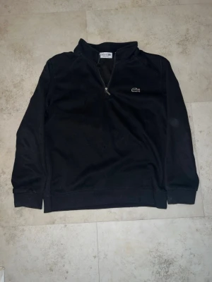 Svart halvzip tröja från Lacoste - Snygg svart tröja från Lacoste med halv dragkedja och klassisk krokodillogo på bröstet. Tröjan har hög krage och långa ärmar, perfekt för en stilren och avslappnad look. Passar lika bra till jeans som till mjukisbyxor.