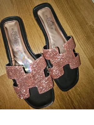 Glittriga sandaler  - Säljer ett par snygga sandaler med breda remmar i rosa glitter och klassisk H-design. Sandalerna har svart sula och öppen tå, samt fyrkantig form framtill. Perfekta för dig som vill sticka ut med lite extra bling.