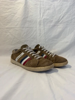 Bruna Moncler sneakers i mocka - Snygga bruna sneakers från Moncler i mocka med vita, röda och blå ränder på sidan. Skorna har vita snören och en beige sula. Klassisk låg modell med Moncler-logga på sidan och tungan. Perfekta för dig som gillar stilrena och sportiga detaljer.