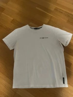 Sail Racing Tröja - Vit t-shirt från Sail Racing med svart logga tryckt på bröstet. Klassisk rund hals och korta ärmar. Tillverkad i mjuk bomull som känns skön mot huden. Perfekt till jeans eller shorts för en clean och enkel stil.