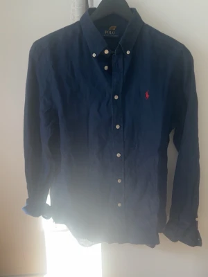 Marinblå skjorta från Polo Ralph Lauren - Klassisk marinblå skjorta från Polo Ralph Lauren med knappar framtill och button-down krage. Skjortan har lång ärm och den ikoniska röda logotypen broderad på bröstet. Tillverkad i linne för en skön  känsla. Dunder till sommaren.priset är diskutabelt.