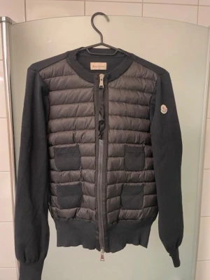 Svart cardiganjacka från Moncler - Snygg svart cardiganjacka från Moncler med quiltad framsida och stickade ärmar samt rygg. Jackan har rund halsringning, dragkedja framtill och fyra dekorativa fickor på framsidan. Moncler-logga på ena ärmen. Perfekt för lager på lager och streetstyle. Storlek är 1 vilket motsvarar S, både NFC och Qr koden funkar 