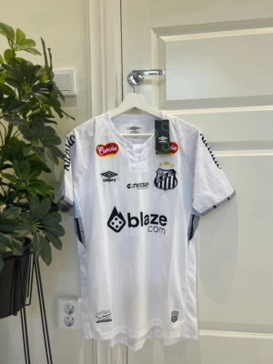 Vit Santos FC matchtröja från Umbro - Säljer en vit matchtröja från Santos FC med svarta detaljer och flera tryckta loggor, bland annat blaze.com och Umbro. Tröjan har korta ärmar, rund hals och klubbmärke på bröstet. Perfekt för fotbollsfans som vill sticka ut på planen eller läktaren.