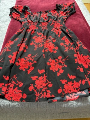 Svart och röd blommig klänning - Supersnygg svart klänning med röda blommor och smock i midjan. Offshoulder-modell med korta puffärmar och volangdetaljer upptill. Perfekt för dig som vill sticka ut med ett färgstarkt mönster och trendig passform.