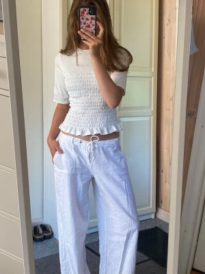 Vit smockad topp med volangkant - Säljer en söt vit topp med smockad midja och volangkant nedtill. Toppen har rund halsringning och halvlånga ärmar. Perfekt till både jeans och kjol för en fräsch look.
