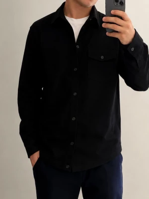 Svart overshirt med knappar - Snygg svart overshirt med klassisk krage och knäppning framtill. Två bröstfickor med lock och knappar ger en clean look. Perfekt att bära över en t-shirt för en avslappnad stil. Tillverkad i mjukt material för extra komfort.
