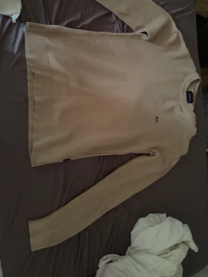 Lindahls tröja  - Snygg beige stickad tröja med rund hals och diskret logga på bröstet. Tröjan har långa ärmar och ribbade muddar vid ärmslut och nederkant. Perfekt för lager på lager och enkel att matcha med olika outfits.