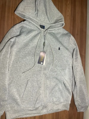 Grå zip hoodie från Polo Ralph Lauren - Snygg ljusgrå hoodie med dragkedja från Polo Ralph Lauren. Klassisk design med huva och dragsnören, två stora fickor framtill och den ikoniska lilla loggan broderad på bröstet. Perfekt till chill vardagsstil och enkel att matcha med allt. Säljer pga av att de inte passar. Skriv vid minsta funderingar. 