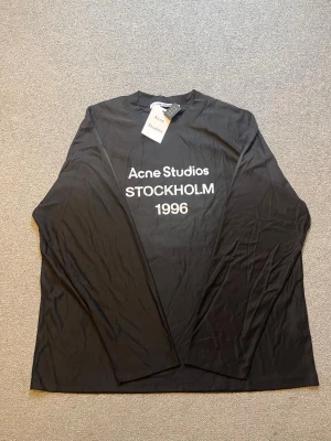 Svart långärmad topp Acne Studios - Svart långärmad topp från Acne Studios med tryck i vitt framtill där det står 'Acne Studios STOCKHOLM 1996'. Klassisk rak passform och rund halsringning. Perfekt för dig som gillar stilrena och trendiga plagg.