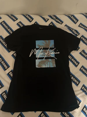 Svart McKenzie t-shirt - Svart t-shirt med blått palmtryck och vit McKenzie-logga på bröstet. Klassisk passform med rund hals och korta ärmar. Perfekt för en avslappnad stil.