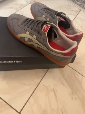 Grå Onitsuka Tiger sneakers - Snygga grå sneakers från Onitsuka Tiger med beigea detaljer och röd insida. Skorna har klassisk låg profil, gummisula och ovandel i mocka och textil. Perfekta för dig som gillar retrostil och vill ha något bekvämt till vardags.