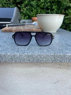 Svarta Tom Ford solglasögon - Snygga svarta solglasögon från Tom Ford med fyrkantiga bågar och mörka tonade glas. Klassisk och stilren design med diskreta gulddetaljer vid gångjärnen. Perfekta för att lyfta din look under soliga dagar.