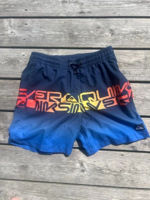 Quiksilver badbyxor med färgskiftning, stl M/12y - Säljer ett par snygga Quiksilver badshorts i blått med cool färgskiftning till rött och gult över låren. Shortsen har elastisk midja med snörning och stort Quiksilver-tryck runt om. Perfekta för stranden eller poolen. Materialet känns som snabbtorkande polyester.