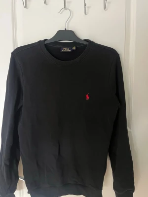 Svart sweatshirt från Polo Ralph Lauren - Klassisk svart sweatshirt från Polo Ralph Lauren med rund halsringning och röd broderad logga på bröstet. Tröjan har ribbade muddar vid ärmslut och nederkant. Perfekt för en clean och stilren look.