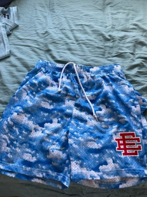 Blå meshshorts med moln och brodyr - Säljer ett par blå meshshorts med molnliknande mönster och vit snörning i midjan. På vänster ben finns en röd och vit broderad logga. Perfekta för sommaren och riktigt sköna att ha på sig.