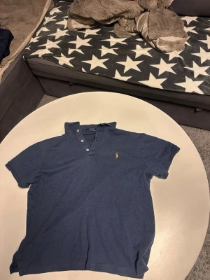 Blå pikétröja från Polo Ralph Lauren - Säljer en klassisk blå pikétröja från Polo Ralph Lauren i modellen Custom Slim Fit. Tröjan har korta ärmar, knappar vid halsen och den ikoniska broderade loggan på bröstet. Perfekt för en avslappnad men ändå stilren look. Kontakta mig om ni har frågor 