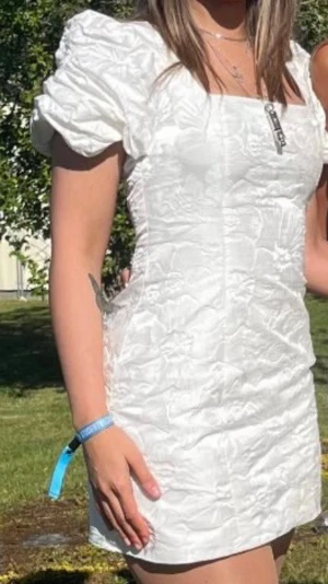Vit kortklänning med puffärm - Vit kortklänning med fyrkantig urringning och puffiga kortärmar. Klänningen har ett diskret blommönster i tyget och en figurnära passform som ger en smickrande siluett. Perfekt som studentklänning eller till sommarens alla festliga tillfällen.  Klänningen är helt ny och passar storlek S–M.