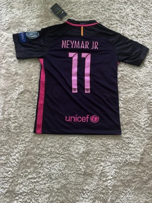 Barça Neymar Jr matchtröja lila/rosa - Snygg FC Barcelona matchtröja från Nike i mörklila med rosa detaljer. Neymar Jr och nummer 11 på ryggen, officiella Champions League-märken på ärmarna och Qatar Airways-logga på bröstet. Tillverkad i lätt och ventilerande material, perfekt för fotboll eller träning.