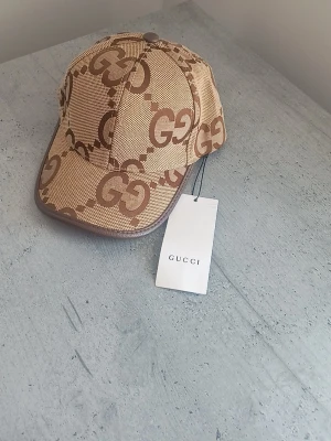Beige keps från Gucci med GG-mönster - Snygg beige keps från Gucci med det klassiska GG-monogrammet i brunt över hela kepsen. Justerbar rem bak i brunt läder och detaljer i samma färg. Tillverkad i Italien och har en modern, sportig look som sticker ut.