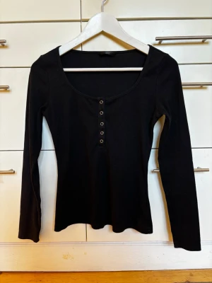 Svart ribbad långärmad topp med djup urringning och knappar  - Svart långärmad topp med ribbad struktur och rundad halsringning. Toppen har knappdetaljer framtill och är figurnära i modellen. Perfekt skick! Polyestertrikå