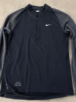 Nike träningströja  - Svart långärmad träningströja från Nike med halv dragkedja framtill och vita detaljer. Ärmarna har ett grått mönster och tröjan är tillverkad i ett lätt, tekniskt material som andas. Perfekt för löpning eller annan träning.