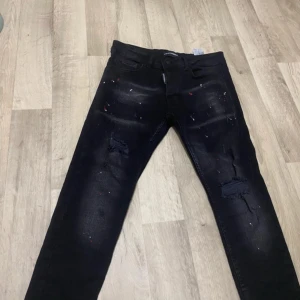 Svarta Dsquared2 jeans med färgstänk - Säljer ett par svarta jeans från Dsquared2 med coola färgstänk i rött och vitt samt slitna detaljer på benen. Jeansen har klassisk femficksdesign, raka ben och är tillverkade i stretchig denim för skön passform.
