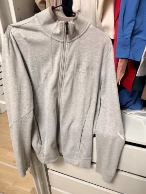 Ljusgrå ziptröja från Hugo Boss - Snygg ljusgrå ziptröja från Hugo Boss med hög krage och diskret logga på bröstet. Regular fit-modell med hel dragkedja och praktiska fickor. Perfekt till både jeans och joggers för en clean och stilren look.