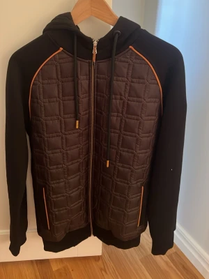 Svart quiltad hoodie Montfleur Paris - Snygg svart hoodie från Montfleur Paris i storlek S. Framsidan är quiltad med rutmönster och har orange detaljer längs dragkedja och fickor. Utrustad med huva och dragsnören. Perfekt för en trendig och avslappnad stil.