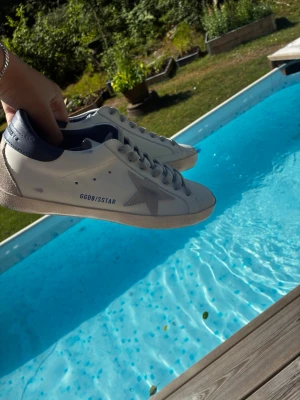 Golden goose skor - Snygga vita sneakers med blå häl och grå stjärndetalj på sidan. Skorna har klassisk låg profil, vita snören och texten GGDB/SSSTAR tryckt på sidan. Perfekta för en stilren och trendig look. Innersulan är 25 och en halv😃