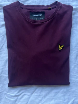 Vinröd t-shirt från Lyle & Scott - Säljer en vinröd t-shirt från Lyle & Scott med klassisk gul logga på bröstet. T-shirten har rund halsringning och korta ärmar. Tillverkad i mjuk bomull som känns skön mot huden. Perfekt för en clean och stilren look.
