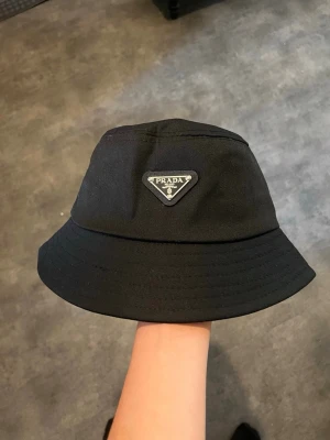 Svart bucket hat från Prada - Säljer en svart bucket hat från Prada med den ikoniska trekantiga loggan framtill. Hatten är tillverkad i ett slitstarkt tyg och har en klassisk form med bred skärm. Perfekt accessoar för en trendig och avslappnad stil.