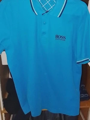 Hugo Boss pikétröja - Snygg blå pikétröja från Hugo Boss med klassisk krage och knappar framtill. Tröjan har vita och mörkblå detaljer på krage och ärmslut samt diskret logga på bröstet. Perfekt för en stilren och sportig look.