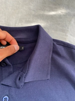 Mörkblå pikétröja med krage - Säljer en stilren mörkblå pikétröja i klassisk modell med krage och knappar framtill. Tröjan är tillverkad i bomull och har en normal passform. Perfekt för en clean och enkel look.