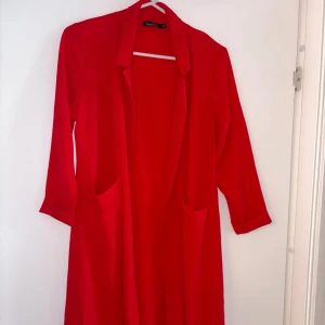 Röd lång kappa från Boohoo - Snygg röd kappa i blus liknande material från Boohoo med lång passform och stora fickor framtill. Jackan har en öppen front utan knäppning och är tillverkad i ett lätt, strukturerat tyg som känns skönt mot huden. Perfekt för dig som vill sticka ut med färg i din outfit. Oanvänd 