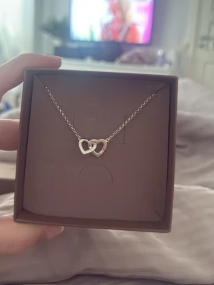 Silverhalsband med dubbla hjärtan - Säljer ett stilrent silverhalsband från AUGUST med två sammanlänkade hjärtan. Kedjan är tunn och smidig, och hjärtana har en modern, minimalistisk design. Perfekt för dig som gillar enkla men snygga smycken.