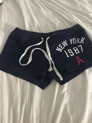 Mörkblå shorts med snörning - Säljer ett par mörkblå shorts med vit snörning i midjan och tryck med texten 'NEW YORK 1987 A' framtill. Shortsen har en sportig look och är perfekta för en helt vanlig dag, från shein, korta men täcker ändå,