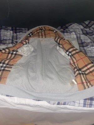 Grå hoodie från Burberry med rutig huva - Snygg grå hoodie från Burberry i storlek M. Utsidan är enkel och stilren medan insidan av huvan har det klassiska Burberry-rutiga mönstret i beige, svart och rött. Perfekt för dig som vill ha en hoodie med exklusiv känsla och ikonisk design.
