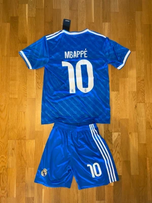 Mbappé Real Madrid bortaställ blå - Säljer ett blått Real Madrid matchställ med Mbappé och nummer 10 på ryggen. Tröjan har vita detaljer, klubbmärke och 'Emirates Fly Better' på bröstet. Shortsen har tre vita ränder och klubbmärke. Perfekt för fotbollsträning eller match.