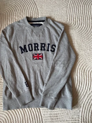 Grå sweatshirt från Morris - Snygg grå sweatshirt från Morris med mörkblå text och en broderad brittisk flagga på bröstet. Tröjan har rund halsringning, ribbade muddar och en avslappnad passform. Perfekt för en casual look.
