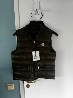 Olivgrön dunväst från Moncler - Snygg olivgrön dunväst från Moncler med quiltad design och hög krage. Västen har två dragkedjeförsedda fickor framtill och en Moncler-logga på bröstet. Tillverkad i glansigt material och stängs med dragkedja. Perfekt för lager-på-lager och streetwear. Helt ny storlek 2