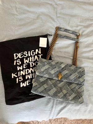 Kurt geiger kensington i denim med guldkedja - Snygg Kurt geiger kensington axelväska i flätad denim med guldfärgad kedja och coolt spänne framtill. Väskan har två vadderade axelband.