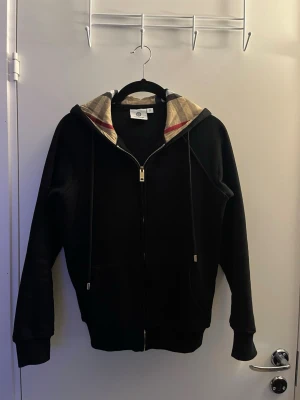 Svart burberry hoodie med mönster för luva och fina detaljer  - Svart hoodie med klassiskt rutigt foder i huvan och dragkedja framtill. Hoodien har snörning i huvan, ribbade muddar och två fickor framtill. Perfekt för en stilren och trendig look.