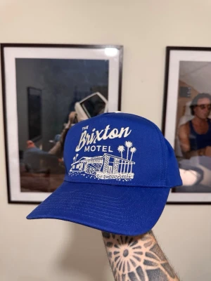 Blå Brixton keps med brodyr - Snygg blå keps från Brixton med vit brodyr framtill där det står 'The Brixton Motel' och en motelldesign. Baksidan har texten 'Clean & Comfortable' broderad. Justerbar snapback och klassisk form. Perfekt för en avslappnad streetstyle.